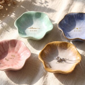 Pu&ograve; includere: Quattro ciotole portagioie in ceramica a forma di petalo, nei colori pastello: verde menta, rosa, blu e beige. Ogni ciotola ha un testo dorato con nomi e date. Una collana d'argento &egrave; appoggiata nella ciotola beige.