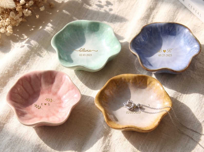 Pu&ograve; includere: Quattro ciotole portagioie in ceramica a forma di petalo, nei colori pastello: verde menta, rosa, blu e beige. Ogni ciotola ha un testo dorato con nomi e date. Una collana d'argento &egrave; appoggiata nella ciotola beige.
