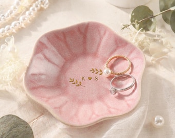 Può includere: Un piatto rosa in ceramica per anelli con un design floreale e scritte dorate "K ♥ S". Due anelli, uno dorato con una perla e uno argentato con un diamante, poggiano sul piatto. Perle e verde circondano il piatto.