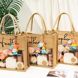 May include: Three burlap tote bags with clear front panels, each personalized with a name in black script. The bags are filled with flowers, gifts, and decorative items. Each bag has a colorful tassel keychain.