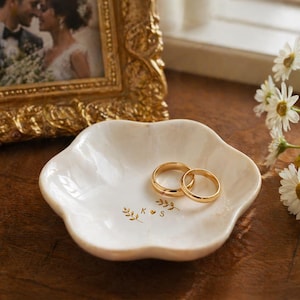 May include: A white, scalloped-edge ring dish with two gold wedding bands. The dish has gold floral accents and the initials "K & S". A framed photo and flowers are in the background.