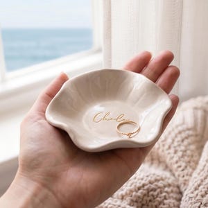 Pu&ograve; includere: Un piccolo portagioie in ceramica bianco sporco con un bordo ondulato, tenuto in mano. Il piatto ha il nome "Chloe" in oro e contiene un anello d'oro. Lo sfondo include una finestra con vista sul mare e una coperta beige lavorata a maglia.