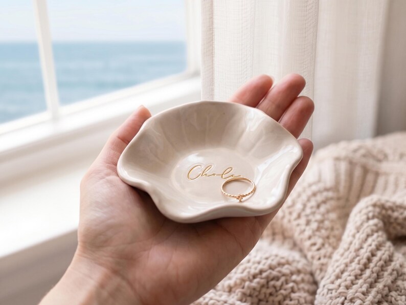 Pu&ograve; includere: Un piccolo portagioie in ceramica bianco sporco con un bordo ondulato, tenuto in mano. Il piatto ha il nome "Chloe" in oro e contiene un anello d'oro. Lo sfondo include una finestra con vista sul mare e una coperta beige lavorata a maglia.