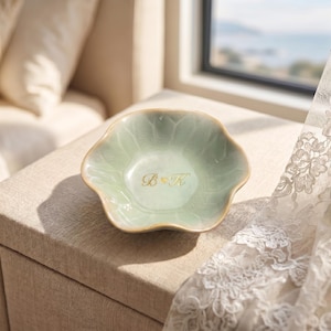 Pu&ograve; includere: Un piatto in ceramica verde chiaro, con bordo smerlato e iniziali dorate "B & K". Il piatto &egrave; su una superficie beige, con una finestra e una tenda di pizzo sullo sfondo. Il piatto &egrave; probabilmente per gioielli o piccoli oggetti.