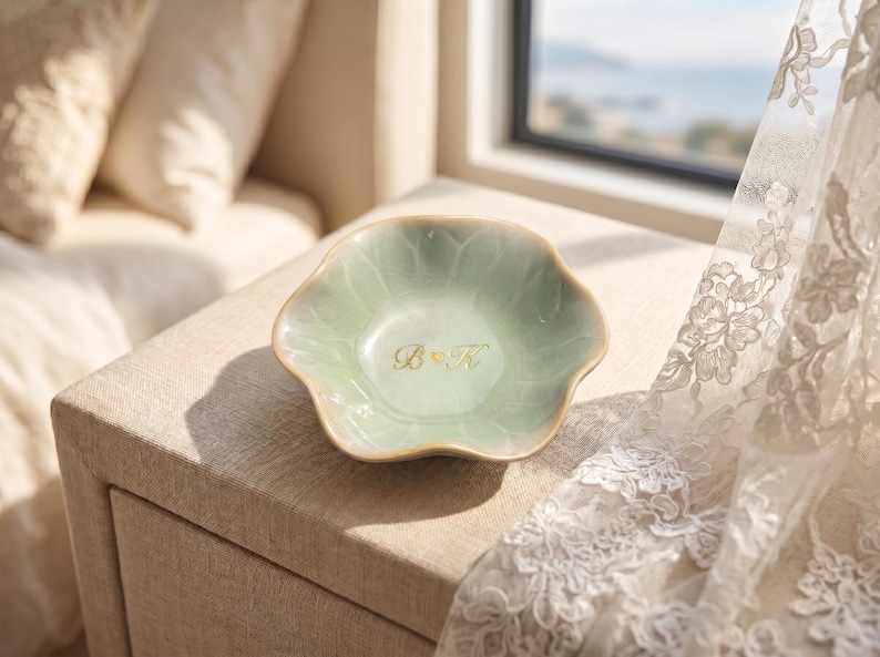 Pu&ograve; includere: Un piatto in ceramica verde chiaro, con bordo smerlato e iniziali dorate "B & K". Il piatto &egrave; su una superficie beige, con una finestra e una tenda di pizzo sullo sfondo. Il piatto &egrave; probabilmente per gioielli o piccoli oggetti.