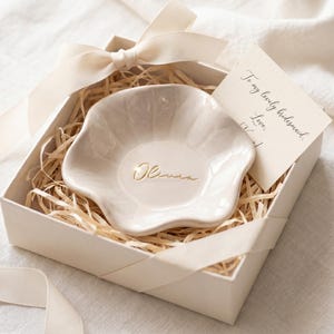 Pu&ograve; includere: Un portagioie in ceramica color crema a forma floreale, in una confezione regalo con paglia e un nastro color crema. Il piatto &egrave; inciso con il nome "Olivia" in oro. Un piccolo biglietto recita "To my lovely bridesmaid."