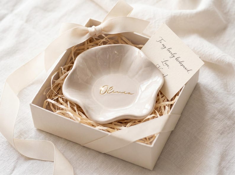 Pu&ograve; includere: Un portagioie in ceramica color crema a forma floreale, in una confezione regalo con paglia e un nastro color crema. Il piatto &egrave; inciso con il nome "Olivia" in oro. Un piccolo biglietto recita "To my lovely bridesmaid."