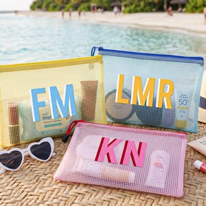 May include: Three rectangular mesh zip pouches in yellow, light blue, and pink. The yellow pouch features the letters FM, the blue pouch features LMR, and the pink pouch features KN. Each pouch contains various personal care items and cosmetics.