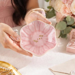 Pu&ograve; includere: Un piccolo piatto in ceramica rosa a forma di petalo. Il piatto presenta un design acquerello rosa chiaro e dettagli di foglie dorate con le lettere "K S". Ideale per contenere gioielli o piccoli oggetti.