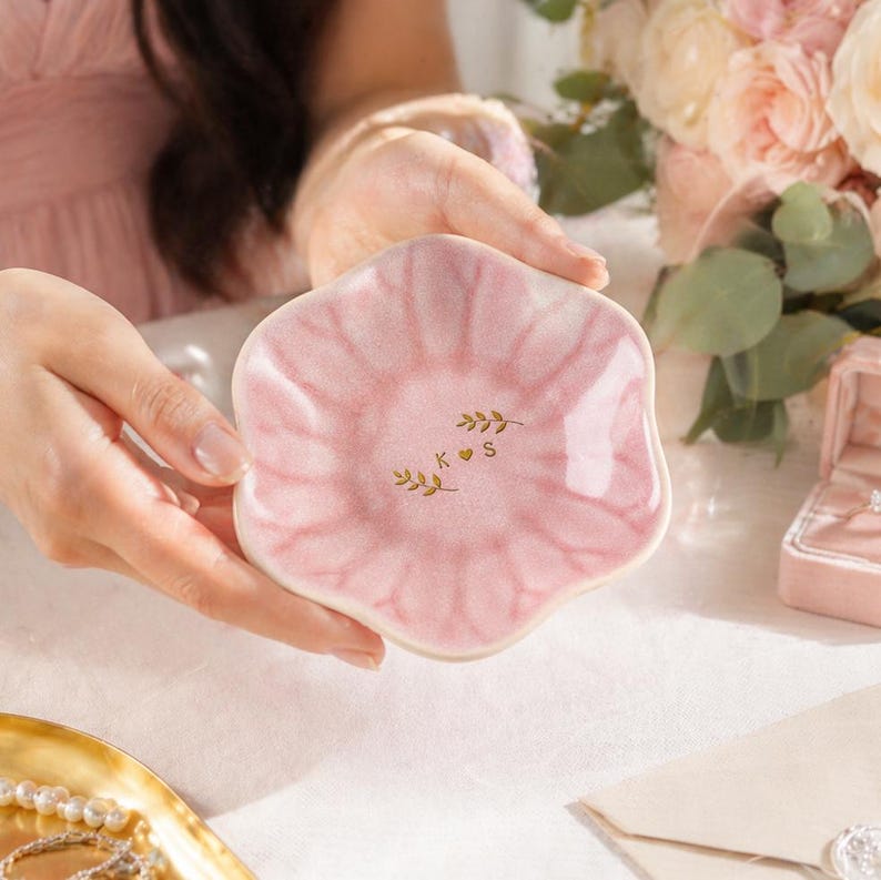Pu&ograve; includere: Un piccolo piatto in ceramica rosa a forma di petalo. Il piatto presenta un design acquerello rosa chiaro e dettagli di foglie dorate con le lettere "K S". Ideale per contenere gioielli o piccoli oggetti.