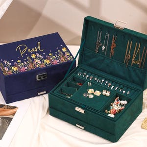 Può includere: Due portagioie. Uno è blu navy con accenti floreali e la parola "Pearl" in corsivo dorato. L'altro è aperto, verde smeraldo e pieno di anelli, collane e orecchini. Entrambi hanno finiture dorate.