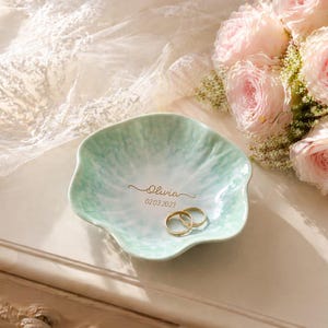 Pu&ograve; includere: Un piatto portaanelli in ceramica verde chiaro, con bordi smerlati, con il nome "Olivia" e la data "02.03.2023" in oro. Due fedi d'oro sono all'interno. Un bouquet di rose rosa &egrave; sullo sfondo.