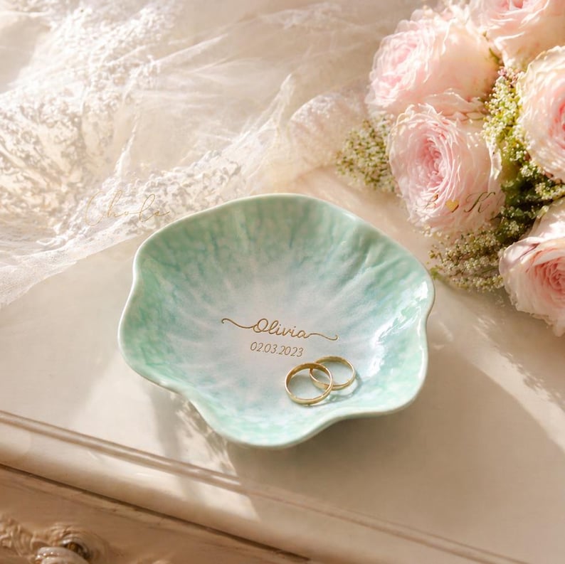 Pu&ograve; includere: Un piatto portaanelli in ceramica verde chiaro, con bordi smerlati, con il nome "Olivia" e la data "02.03.2023" in oro. Due fedi d'oro sono all'interno. Un bouquet di rose rosa &egrave; sullo sfondo.