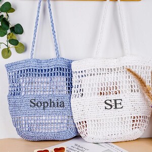May include: Two handmade crochet tote bags, one light blue and one white, with braided handles. The light blue bag has the name "Sophia" printed on it, and the white bag has the initials "SE".