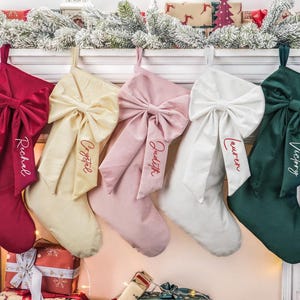 Pode incluir: Cinco meias de Natal de veludo em vermelho, creme, rosa, branco e verde, cada uma com um grande laço e um nome bordado na frente. As meias estão penduradas numa lareira decorada com vegetação e presentes.