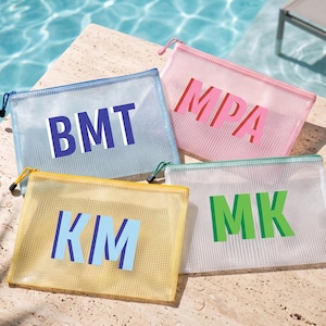 May include: Four rectangular mesh zipper pouches in light blue, pink, yellow, and clear. Each pouch features bold, block-letter monograms in contrasting colors: BMT in dark blue, MPA in red, KM in light blue, and MK in green. Each has a zipper closure.