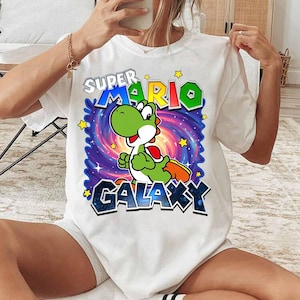 May include: White t-shirt featuring a colorful graphic of Yoshi from Super Mario Galaxy. The design includes the text "SUPER MARIO GALAXY" with a cartoon Yoshi character set against a swirling galaxy background. Yellow stars are scattered around the design.