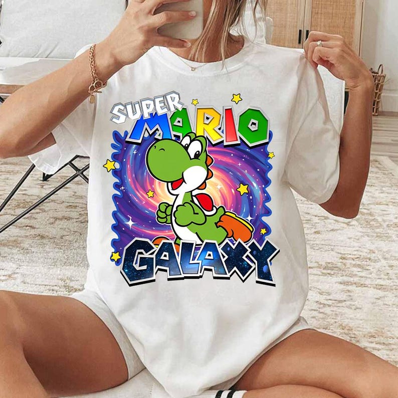 May include: White t-shirt featuring a colorful graphic of Yoshi from Super Mario Galaxy. The design includes the text "SUPER MARIO GALAXY" with a cartoon Yoshi character set against a swirling galaxy background. Yellow stars are scattered around the design.