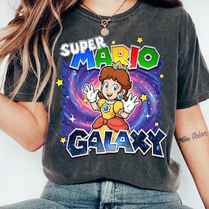 May include: Dark gray t-shirt featuring a colorful graphic of Princess Daisy from Super Mario Galaxy. The design includes the text "SUPER MARIO GALAXY" with a galaxy background and stars. The princess is wearing a yellow dress.