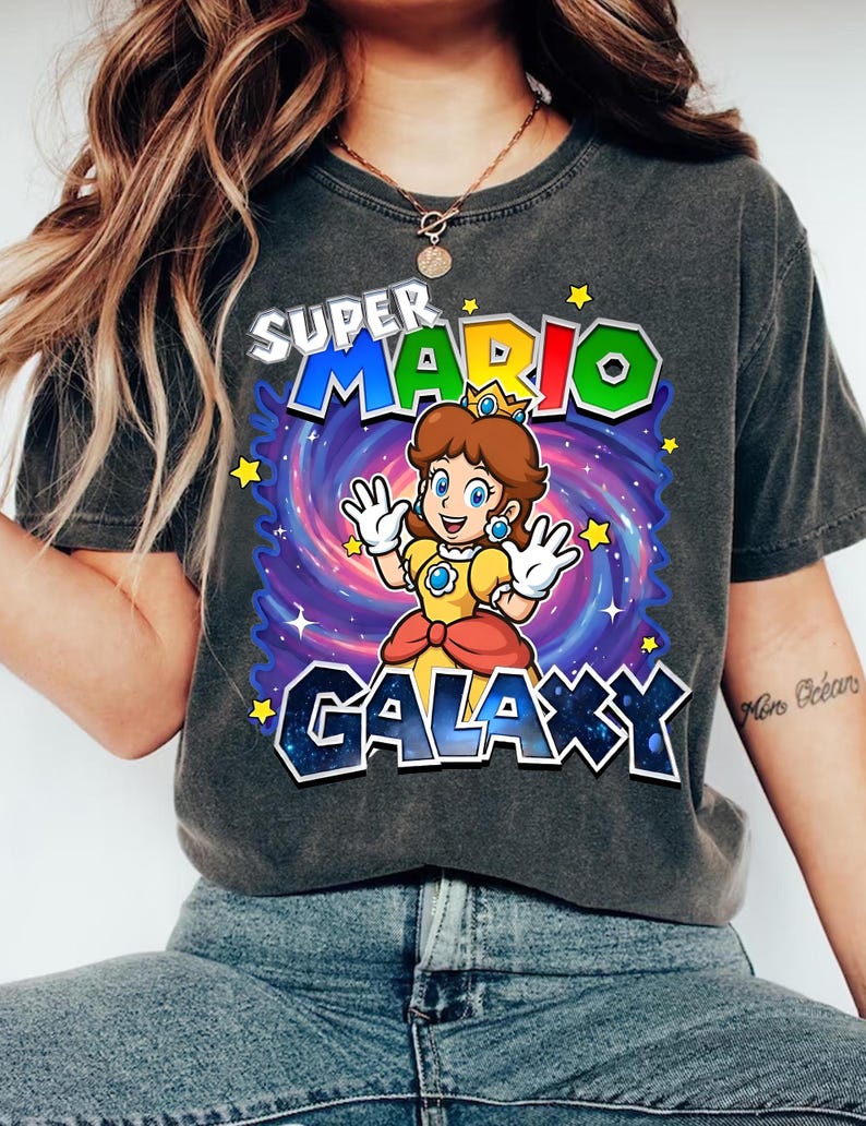May include: Dark gray t-shirt featuring a colorful graphic of Princess Daisy from Super Mario Galaxy. The design includes the text "SUPER MARIO GALAXY" with a galaxy background and stars. The princess is wearing a yellow dress.