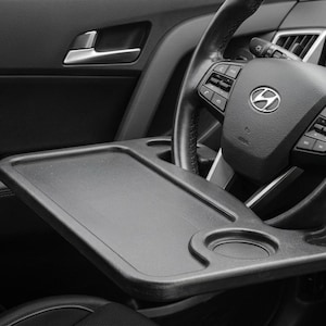 May include: A black plastic steering wheel tray featuring a flat rectangular surface and a circular cup holder, mounted horizontally across the center of a black Hyundai steering wheel within the interior of a vehicle.