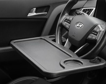 May include: A black plastic steering wheel tray featuring a flat rectangular surface and a circular cup holder, mounted horizontally across the center of a black Hyundai steering wheel within the interior of a vehicle.
