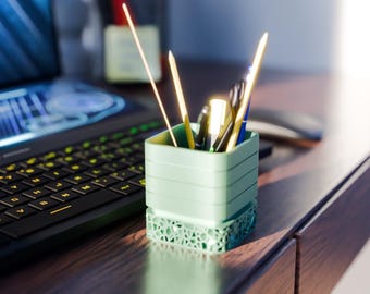 May include: A light green desk organizer filled with pens, pencils, and other office supplies sits on a wooden desk. The organizer has a geometric patterned base and a square shape. A laptop is visible in the background.