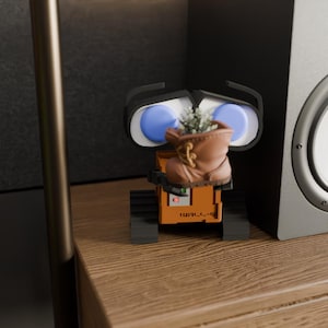May include: A detailed, orange WALL-E figurine with blue eyes and a brown boot planter filled with greenery. The figurine is on a wooden surface, with a speaker and a metal pole in the background.