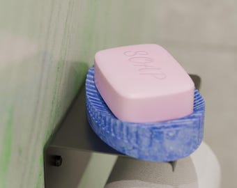 May include: A pink bar of soap with the word "SOAP" embossed on top rests in a blue soap dish. The soap dish is on a metal shelf, with a roll of toilet paper below. The background is a light green and white wall.