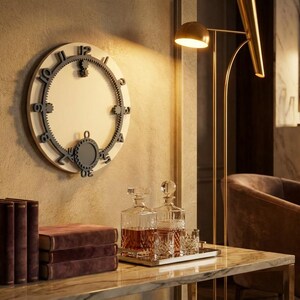 May include: A circular wall clock with a white face and a dark gray gear-shaped outer ring featuring raised numbers. The clock has a small circular pendulum at the bottom and is mounted on a textured wall above a table with books and glassware.
