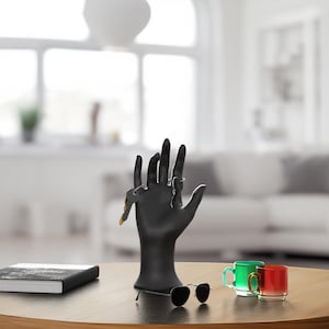 Op de afbeelding: Een zwarte handvormige sieradenstandaard met ringen. Een zonnebril, een gesloten boek en twee kleine glazen mokken staan op een ronde houten tafel. De mokken zijn groen en rood.