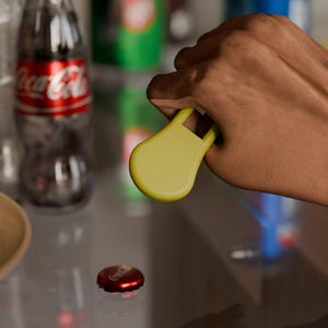 May include: A yellow bottle opener is being used to remove a red Coca-Cola bottle cap. The opener has a rounded shape with a handle. A Coca-Cola bottle is visible in the background.