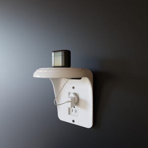 Könnte beinhalten: Ein weißes, an der Wand befestigtes Regal mit integrierter Steckdose. Auf dem Regal steht eine kleine, dunkle Box. Ein weißer Stecker ist in die Steckdose gesteckt, mit einem Kabel, das davon abgeht. Das Regal ist an einer dunkelgrauen Wand befestigt.