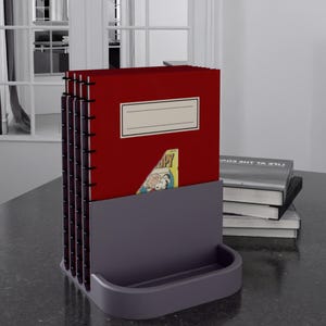 May include: A dark grey desk organiser holding several red notebooks with black spiral bindings. A comic book page is visible in one notebook. A small tray is at the base of the organiser. A stack of books is to the right.