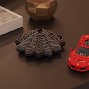 Peut inclure: Un objet géométrique gris foncé et marron avec un trou central, ressemblant à une fleur ou une étoile stylisée. Une voiture jouet rouge est visible en arrière-plan, ainsi qu'un bol décoratif de couleur or.