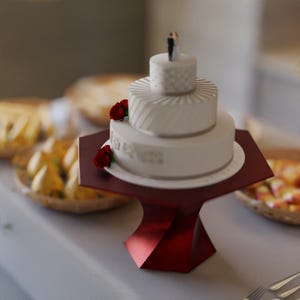 May include: A three-tiered white wedding cake with silver trim and red rose accents, topped with a bride and groom figurine. The cake sits on a red, geometric cake stand. The cake is approximately 6 inches tall.