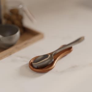 Può includere: Un cucchiaio in acciaio inossidabile poggia su un piccolo poggia cucchiaio in legno. Il poggia cucchiaio è di colore marrone caldo e ha una forma curva. Lo sfondo è una scena di cucina sfocata con un vassoio e un barattolo di legno.