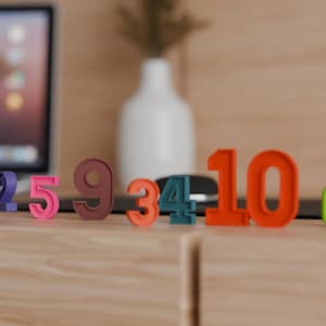 Puede incluir: Una colección de números tridimensionales coloridos del 1 al 10, en varios tonos de rosa, naranja, verde, azul y marrón, están dispuestos sobre una superficie de madera. Un portátil y un jarrón blanco son visibles en el fondo.