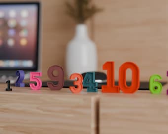 May include: A collection of colorful, three-dimensional numbers from 1 to 10, in various shades of pink, orange, green, blue, and brown, are arranged on a wooden surface. A laptop and a white vase are visible in the background.