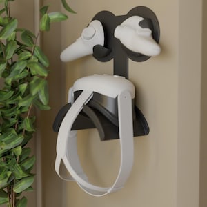 Puede incluir: Un soporte de pared para auriculares de realidad virtual en blanco y negro. El soporte sujeta los auriculares y los mandos. Los auriculares son blancos y negros, con una correa blanca. Los mandos son blancos. El soporte está fijado a una pared beige.