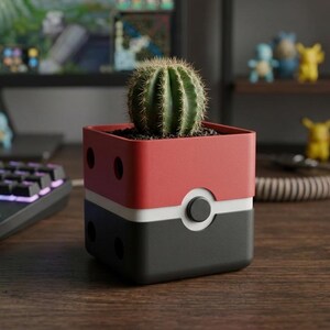 May include: A square-shaped planter featuring a red top half and a black bottom half, separated by a white horizontal band with a central circular button detail. A small green cactus is planted inside the container, which has small circular holes.