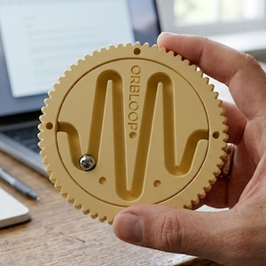 May include: A circular, pale yellow gear-shaped puzzle toy featuring a central, vertical, wave-like track with the word ORBLOOP embossed on the surface. A small, silver-colored metal ball sits within the track, which is designed for movement.
