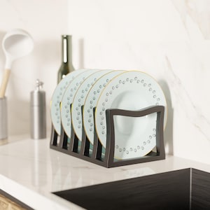 Puede incluir: Un estante de metal marrón oscuro que sostiene cinco platos blancos con un borde azul claro y ribete dorado. Los platos tienen un patrón decorativo de pequeños puntos grises. El estante está sobre una encimera blanca.
