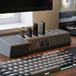 May include: A dark gray rectangular storage organizer for electronic accessories featuring multiple vertical slots for USB flash drives and horizontal slots for SD memory cards, positioned on a desk surface near a computer keyboard and mouse.