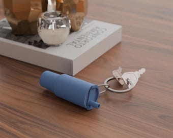 May include: A blue keychain with a cylindrical attachment and two keys. The keychain is on a wooden surface, with decorative gold-colored geometric vases and a book in the background.