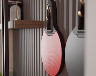 May include: A red and black oval-shaped object with a textured surface, hanging on a vertical, light-colored, slatted wall. The object has a black handle with gold accents. Another similar object is partially visible to the right.