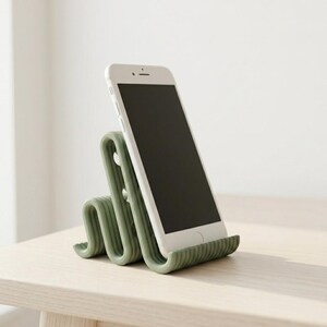 Peut inclure: Un support pour smartphone vert sauge au design ondulé et sinueux. Le support présente une texture nervurée et maintient un smartphone blanc en position verticale inclinée. La structure est composée de boucles continues et répétitives.