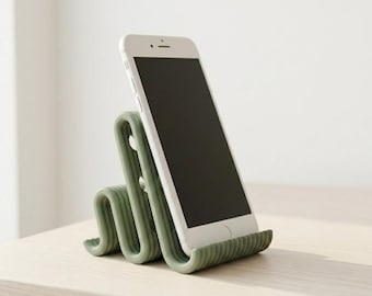 May include: A sage green smartphone stand with a wavy, serpentine design. The stand features a ribbed texture and holds a white smartphone in an upright, angled position. The structure is composed of continuous, repeating loops that provide support.