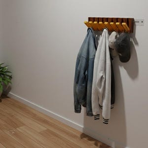 May include: A wooden coat rack fixed to a white wall, featuring several yellow folding hooks. Three jackets and a baseball cap are suspended from the hooks. The floor is wooden.