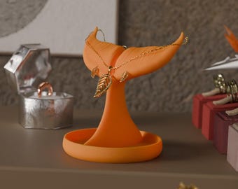 May include: An orange jewelry holder shaped like a whale tail, displaying a gold leaf pendant necklace and earrings. The holder has a circular base and a vertical support. A silver ring box and other jewelry are in the background.